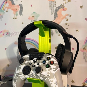 May include: A green 3D printed stand holds a black gaming headset and a white camouflage Xbox controller with the word "RECON" printed on it.