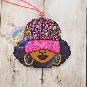 May include: A glittery black, pink, and gold ornament featuring a stylized face with a pink headband, gold hoop earrings, and dark hair. The ornament has a string for hanging and a logo that says "Mee Mee's Creations".