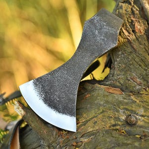 May include: A forged steel axe head with a textured, dark gray finish and a polished silver blade. The axe is resting on a piece of wood, showcasing its craftsmanship and design. The axe is a tool for cutting wood.