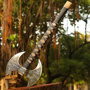 May include: A double-headed axe with a black leather wrapped handle and intricate metalwork. The axe is resting on a wooden stump with a blurred background of green trees.