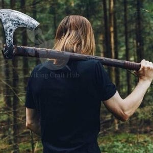 Puede incluir: Un hacha grande y desgastada con una hoja plateada y un mango de madera oscura. El hacha se lleva sobre el hombro. El mango está envuelto en un material marrón oscuro. El hacha es un arma de estilo vikingo.