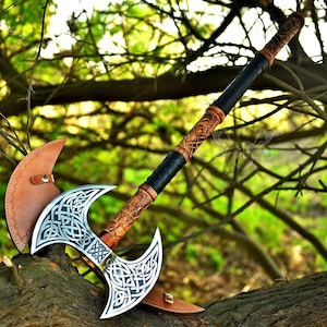 May include: A double-headed axe with a brown leather wrapped handle and a silver blade with intricate Celtic knotwork designs. The axe is resting on a tree branch.