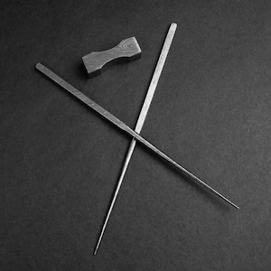May include: Two silver chopsticks crossed over each other on a black surface. A small silver chopstick rest is also on the surface.