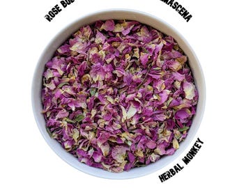 Op de afbeelding: Een witte kom gevuld met gedroogde roze rozenblaadjes. De tekst "ROSE BUD PETALS PINK - ROSA DAMASCENA" staat in het zwart boven de kom en "HERBAL MONKEY" staat in het zwart onder de kom.