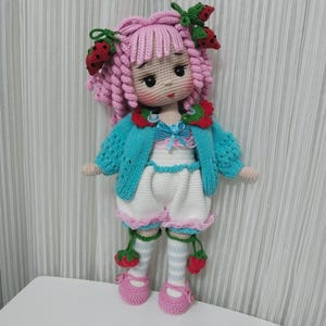 May include: A handmade crochet doll with pink curly hair, adorned with strawberry accents. The doll wears a turquoise cardigan, white shorts, and pink shoes. The doll's outfit includes a blue bow and strawberry details.