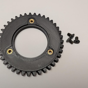 Könnte beinhalten: Ein schwarzes Zahnrad mit Zähnen, mit drei Messing-Einsätzen und drei kleinen schwarzen Schrauben. Das Zahnrad hat eine zentrale kreisförmige Öffnung. Dieses mechanische Bauteil wird wahrscheinlich in Maschinen oder technischen Anwendungen verwendet.