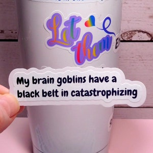 May include: White tumbler with the text "My brain goblins have a black belt in catastrophizing." A hand holds a sticker with the same text. Additional stickers are scattered on a pink surface.