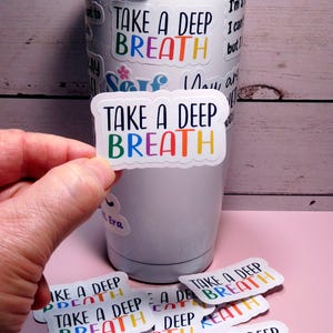 May include: A white tumbler with multiple colorful stickers that say "Take a Deep Breath". The stickers have a rainbow color scheme and a white outline.