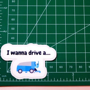 May include: A white sticker with a blue and gray cartoon illustration of a snow plow truck. The sticker says "I wanna drive a..."