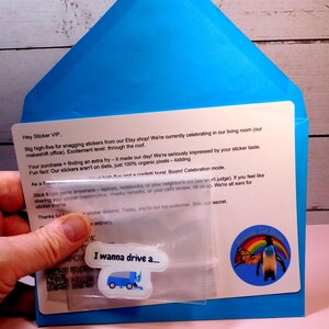May include: A blue envelope with a white card inside. The card has a message thanking a customer for their purchase and includes a small plastic bag with a sticker inside. The sticker features a blue garbage truck with the text "I wanna drive a..." The envelope also has a sticker with a penguin and a rainbow.