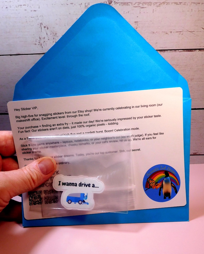 May include: A blue envelope with a white card inside. The card has a message thanking a customer for their purchase and includes a small plastic bag with a sticker inside. The sticker features a blue garbage truck with the text "I wanna drive a..." The envelope also has a sticker with a penguin and a rainbow.
