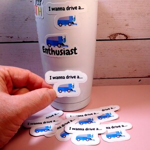 May include: A white tumbler with a blue and white sticker that says "I wanna drive a..." and a blue and white illustration of a snowplow. The tumbler is surrounded by several more of the same stickers.