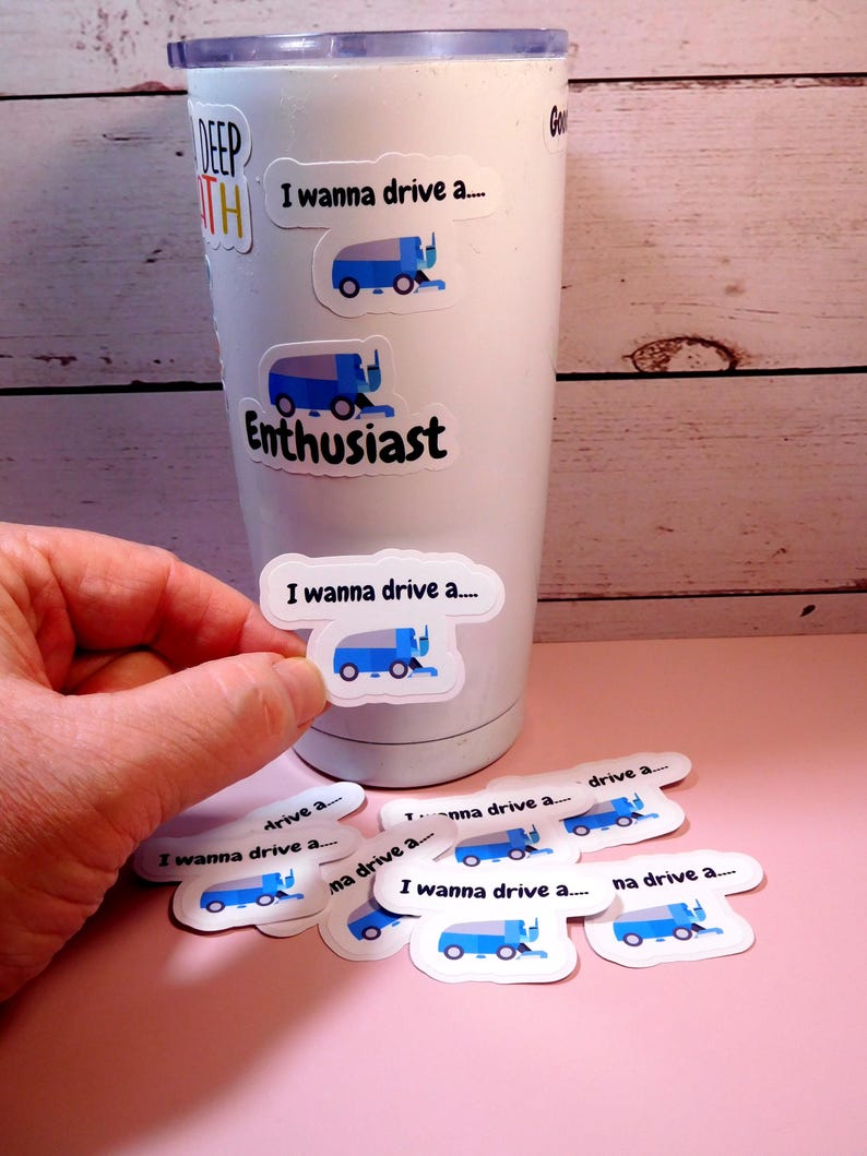 May include: A white tumbler with a blue and white sticker that says "I wanna drive a..." and a blue and white illustration of a snowplow. The tumbler is surrounded by several more of the same stickers.