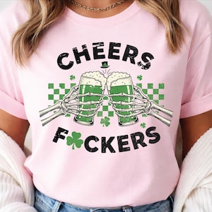 May include: Pink t-shirt with a St. Patrick's Day graphic. The design features skeleton hands toasting green beer, the words "CHEERS F*CKERS", shamrocks, and a green and white checkered pattern.