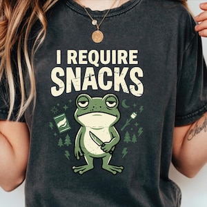 May include: Dark gray t-shirt with the text "I REQUIRE SNACKS" above a green frog holding a knife. The frog is surrounded by illustrations of snacks, trees, and lightning bolts. The shirt is a casual wear item.