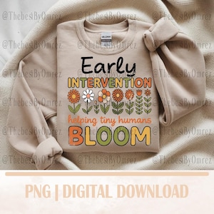May include: A beige sweatshirt with the words "Early Intervention" in black and orange, with a floral design. Below the text is "helping tiny humans BLOOM" in orange and green. The sweatshirt has long sleeves.