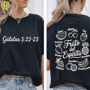 May include: Black t-shirt with white text "Gálatas 5:22-23" on the front. The back features a design with the words "Fruto del Espíritu" and illustrations of fruits like watermelon, cherries, and a pineapple.