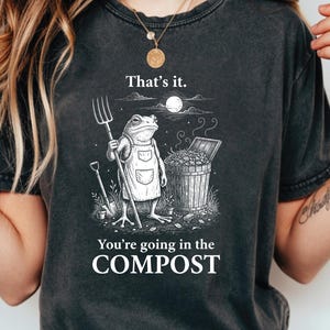 May include: Dark gray t-shirt featuring a whimsical illustration of a frog in an apron holding a pitchfork, standing near a compost bin. The text "That's it. You're going in the COMPOST" is printed below the illustration. The design is in black and white.