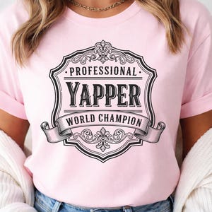 Könnte beinhalten: Hellrosa T-Shirt mit einem schwarz-weißen Grafikdesign. Der Aufdruck lautet "PROFESSIONAL YAPPER WORLD CHAMPION" in einem dekorativen Schild und Banner. Die Ärmel sind hochgekrempelt.
