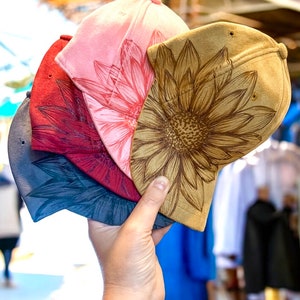 May include: Four different colored baseball caps with sunflower designs. The caps are stacked on top of each other and are being held by a hand.