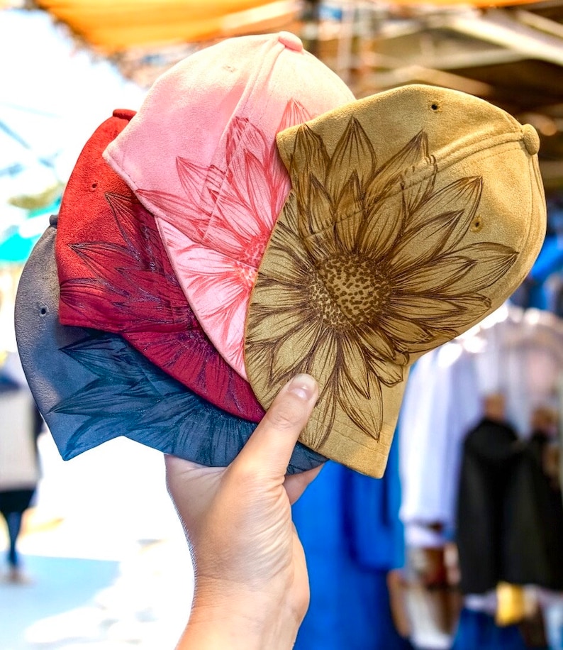 May include: Four different colored baseball caps with sunflower designs. The caps are stacked on top of each other and are being held by a hand.