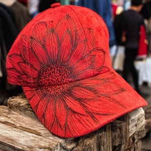 May include: Red baseball cap with a black sunflower design. The cap is made of a soft, suede-like material.