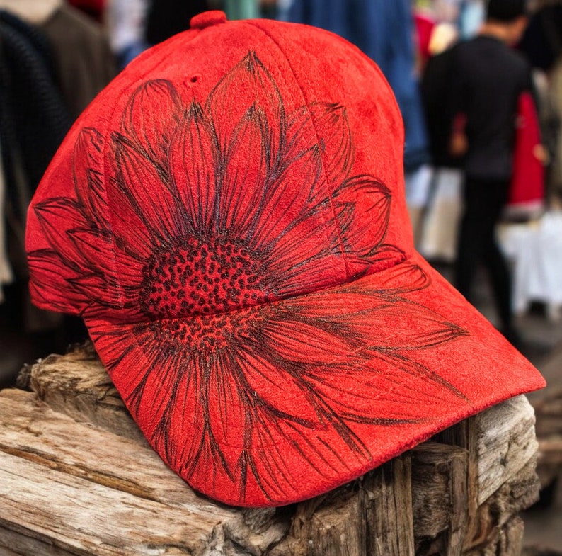 May include: Red baseball cap with a black sunflower design. The cap is made of a soft, suede-like material.