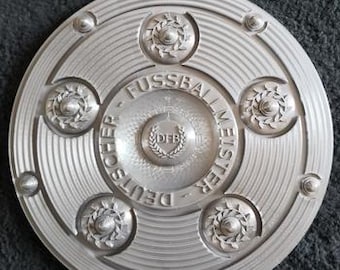 May include: A silver-colored, circular emblem with the words "DEUTSCHER FUSSBALL MEISTER" and "DFB" in the center. The emblem has a series of concentric circles and decorative floral elements. The background is a dark gray.