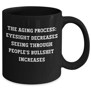 May include: Black ceramic mug with white text: "THE AGING PROCESS: EYESIGHT DECREASES SEEING THROUGH PEOPLE'S BULLSHIT INCREASES." The mug has a large handle and a glossy finish. Ideal for tea or coffee.