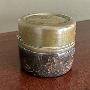 May include: A small, antique round container with a brass lid and a dark brown, textured leather body. The lid is slightly domed and the leather shows signs of age and wear, suggesting it is vintage.