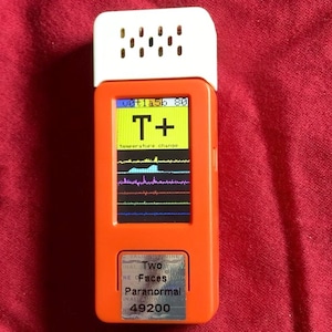Puede incluir: Dispositivo de mano naranja y blanco con una pantalla negra que muestra una gráfica y el texto "T+" y "cambio de temperatura". El dispositivo tiene una pegatina en la parte inferior que dice "Two Faces Paranormal 49200".
