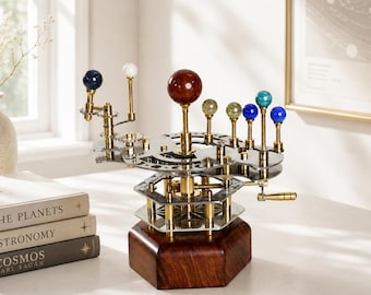 Op de afbeelding: Een mechanisch orrery-model met een centrale donkerrode bol die de zon voorstelt, omringd door kleinere bollen in verschillende kleuren en maten op messing armen, gemonteerd op een zeshoekige houten voet met metalen tandwielen.