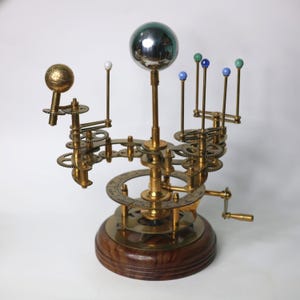 May include: A brass and wood model of the solar system, featuring a central sphere and smaller colored spheres representing planets. The model includes gears, a hand crank, and a wooden base, showcasing intricate mechanical details.