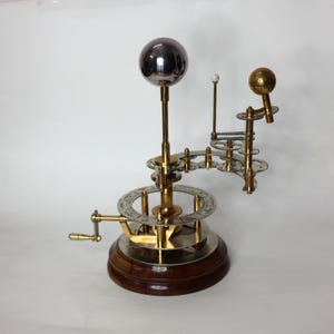 May include: A brass and silver-toned model of the solar system with a hand crank. The model features a central sun, a silver-toned moon, and a gold-toned planet. The model is mounted on a wooden base.