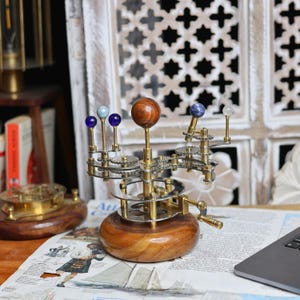 May include: A brass and wood model of the solar system with a wooden base. The planets are represented by small spheres of different colors, including blue, brown, and white. The model is on a table with a newspaper and a laptop computer.
