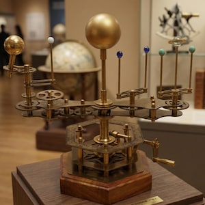 May include: A brass model of the solar system with a large golden sphere at the center and smaller spheres representing planets. The model features gears, levers, and a wooden base, showcasing a detailed mechanical design. The model is displayed in a museum setting.