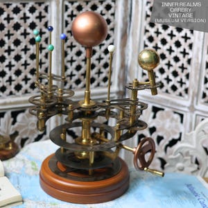May include: A vintage brass orrery model with a copper-colored sun and painted planets. The orrery is on a wooden base and has a crank to rotate the planets.