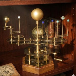 May include: A brass-colored, mechanical orrery model of the solar system. The model features a large central sphere, smaller spheres representing planets, and intricate gears. It is mounted on a wooden base and displayed on a shelf with books.
