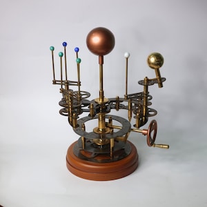 May include: A brass and copper model of the solar system with a wooden base. The model features a central sun with planets orbiting around it. The planets are represented by small colored spheres.