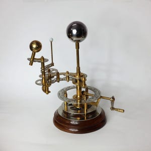 May include: A brass and silver model of the solar system with a wooden base. The model features a sun, earth, and moon, and a crank to rotate the planets.