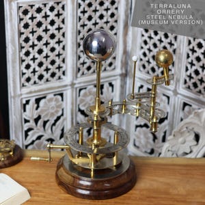 May include: A brass and silver Terraluna orrery with a silver sphere representing the sun, a smaller white sphere representing the moon, and a gold sphere representing a planet. The orrery is on a wooden base and has a white lattice screen in the background. The text "Terraluna Orrery Steel Nebula (Museum Version)" is displayed above the orrery.