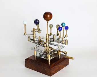 Puede incluir: Un modelo mecánico de orrery que presenta una gran esfera central de madera que representa el sol, rodeada por esferas más pequeñas de varios colores y materiales sobre brazos de latón. El dispositivo incluye engranajes metálicos y una manivela.