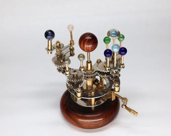 May include: A detailed model of the solar system, featuring a wooden base and a complex arrangement of gears and spheres. The spheres are in various colors, including blue, green, and clear, and are mounted on brass rods. The base is a dark brown color.