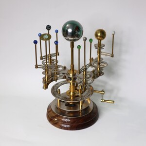 Op de afbeelding: Een messing en zilveren model van het zonnestelsel met een handkruk. Het model heeft een centrale zon met acht planeten die eromheen draaien. De planeten worden weergegeven door kleine gekleurde glazen bollen.