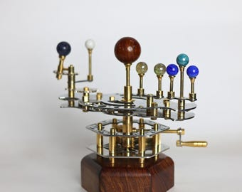 Puede incluir: Un modelo mecánico de orrery que presenta una esfera central de madera que representa el sol, rodeada por esferas más pequeñas de varios colores y materiales montadas sobre brazos de latón y placas de metal plateado, sobre una base de madera.