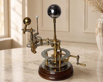 May include: A brass and metal orrery model featuring a large central sphere, a smaller gold-toned sphere, and a tiny white bead on a rod. The mechanism includes interconnected gears, a circular calendar dial, and a hand crank on a wooden base.