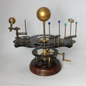 May include: A black and gold metal solar system model with a crank to rotate the planets. The planets are represented by small spheres in various colors, including gold, white, blue, and green.