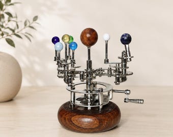 Może przedstawiać: Mechaniczny model orrery z centralną kulą z ciemnego drewna, otoczoną mniejszymi kulkami w kolorze niebieskim, żółtym, zielonym, białym i marmurkowym błękicie, zamontowany na srebrnym metalowym systemie przekładni z drewnianą podstawą i korbką.