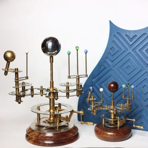 May include: Two brass and wood orrery models of the solar system. The larger model has a silver sphere at its center, with colored spheres representing planets. The smaller model has a wooden sphere at its center. Both have gears and cranks.
