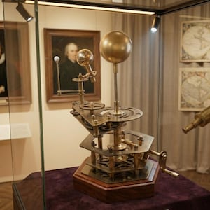 May include: A brass orrery model with a large golden sphere at the top, representing the sun. The model has gears, smaller spheres, and a wooden base. It is displayed in a glass case, with paintings in the background.
